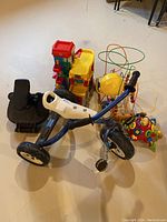 Full view of lot showing tricycle, child seat attachment, and retro toys grouped together on floor with wall in background.
