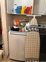 Full view of laundry area showing ironing board, drying rack, washing machine top with assorted detergents and liquids in the background.