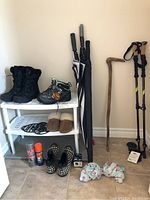 Overview of two pairs of women's boots on a white plastic boot rack, assorted umbrellas, trekking poles, hiking stick, shoe care products, slippers, and boot accessories arranged on tiled floor.