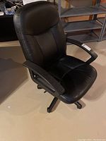 Front and side view of the black office chair showing padded seat, backrest, armrests, and five caster wheels on the base. Minor wear visible on leather.