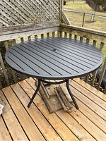 Full view of round black metal patio table on wooden deck with metal slatted top and central umbrella hole.