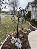 Front view of the black metal pin wheel sculpture mounted in a garden bed with leafless trees and homes in background.