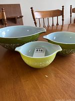 Set of three green Pyrex mixing bowls with white interior and daisy floral pattern on the exterior, displayed on wood dining table.
