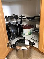 Corner kitchen cabinet showing stacked non-stick frying pans with black handles, a large stainless steel stockpot with lid, a red floral enamel roasting pan with lid, glass rectangular baking dish, and metal roasting rack.