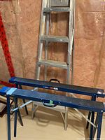 Photo showing the pair of blue Mastercraft filing sawhorses with black tops and the 5-step aluminum ladder against an unfinished basement wall.