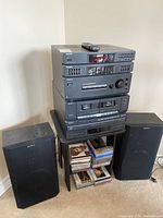 Full view of Sony stereo system stacked with components on small black table flanked by two speakers. CD cases stored on lower shelves of table. Remote control placed on top of stereo.
