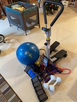 Photo shows the manual stepper with black pedals, blue exercise ball, assorted dumbbells, resistance bands, workout DVDs, and a badminton racket arranged on floor.
