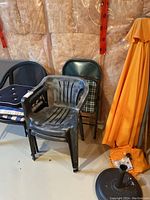 Photo showing four patio chairs of different styles grouped against wall including black plastic chairs and green metal folding chairs with plaid cushions. Orange umbrella and stand in background.