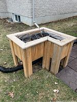Front left-side view of the L-shaped wooden outdoor planter with two black plastic pots inside and visible weathering on wood.