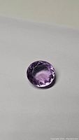 Close-up photo of the round faceted pink-purple amethyst gemstone on a white background showing color and cut details.