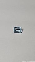 Photo of a natural sky blue topaz gemstone, emerald cut, showing light blue color and clarity on a plain background.