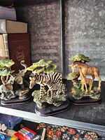 Photo of three animal figurines including zebra with foal, giraffe with calf, and elephant with calf on decorative bases with foliage.
