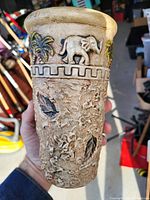 Photo of a tall ceramic vase showing embossed elephant and palm tree design around upper rim and textured leaf motifs on body.