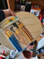 Photo shows a variety of paintbrushes laid out on top of a segmented wooden circular palette or board. The brushes vary in shape and size, including a packaged set of 25 watercolor brushes with blue handles and other loose brushes.