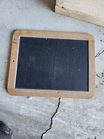 Small chalk board with wooden frame lying on a cracked concrete floor, showing minor use marks, and a hole for hanging at the top center of frame.