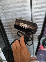 Close-up photo of a hand holding a black Conair hair dryer with '1875' printed on it, showing the body and power cord.