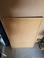 Photo of a large rectangular corkboard with a simple wooden frame leaning against a wall on the floor.