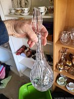 Hand holding a clear glass decanter with a ridged, faceted stopper. Background includes shelves with other glassware and decorative items.