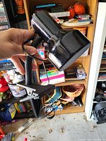 Hand holding a black desk lamp with a flexible neck and clamp base, with attached power cord visible. Background includes shelves and household items.