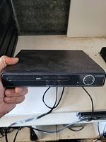 Front view of the black DVD player being held, showing front-loading disc tray, control buttons, and connected cables.