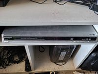 Front view of Panasonic DVD player on a white shelf with buttons and disc tray visible, remote control positioned nearby.