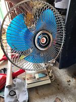 Photo of a blue blade table fan with metal grille and white base, showing the fan's design and working condition.