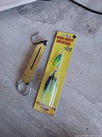 Photo of a metal fishing scale with a hook and ring, next to a new Bird-Shot Boctail spinnerbait fishing lure in original packaging on concrete floor.