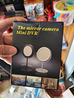 Photo of the packaging for The Mirror Camera Mini DVR showing product in black and white versions with sample room views below