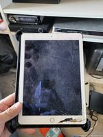 Front view of an iPad showing a white bezel with a round home button, screen heavily cracked and damaged with visible wear and scratches.