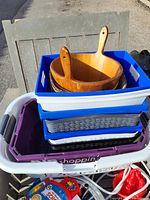 Stack of various plastic storage bins containing wooden kitchenware items on top, photographed outdoors.