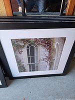 Framed photo print showing an arched window with white wooden frame surrounded by climbing ivy plants. The print is matted in white and framed in black.