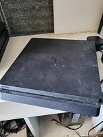 Top view of a black Sony PlayStation 4 console showing dust and wear, placed on a white surface with no cables visible.