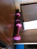 Photo shows two pink dumbbell weights placed on a wooden surface near a cardboard box.