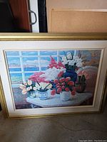 Framed floral still life painting showing flowers in vases on a table with a scenic window view of the ocean and sky.