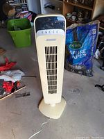 Front view of white Lasko stand up tower fan on a garage floor, surrounded by household items.