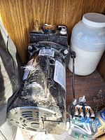 Black electric well pump motor with attached control box and electrical components on a wooden shelf, accompanied by wiring parts and white container.