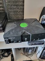 Black original Xbox console on shelf, dusty and signs of wear visible. Front side with controller ports and power button shown.