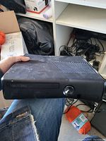 Black Xbox 360 console held in a hand, shown from front and top angle. Dust and wear visible on casing.