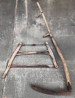 Both the antique crosscut bucksaw and wood handled scythe laid out on a concrete surface showing condition of items, rusted metal blades and aged wooden handles.