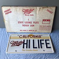 Original Miller High Life cardboard box with shipping labels and text indicating it contains a State License Plate Tacker Sign. Box shows age wear and staining.