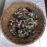 Wide-angle top-down photo showing a large handmade wicker basket filled with hundreds of assorted vintage buttons of various colors, shapes, and sizes, placed on a white surface.