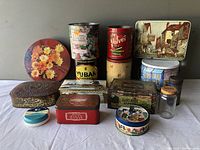 Photo showing assortment of circa 13 vintage tins and 1 small glass jar. Various brands, shapes, colors, and sizes present. Some tins have floral or scenic printed graphics; some brand tins include Velvet Tobacco, Yuban Coffee, Band-Maid Cigars, Stroman's Cigarettes.