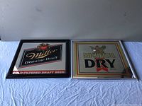 Two framed beer wall signs on a white sheet, Miller Genuine Draft on left, Michelob Dry Beer on right