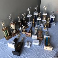 Photo showing 22 assorted vintage trophies and parts on a blue cloth, primarily tennis figures on bases of wood, marble, and plastic, with visible wear and tarnish.