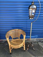 Full view of woven chair and tall metal lantern hanger with hanging lantern in front of a blue corrugated backdrop.