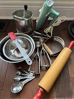 Group photo showing turquoise vintage Sunbeam hand mixer with metal beaters, stainless steel bowls, cookie scoops, measuring spoons, rolling pin, sifter, pastry blender, and silicone spatula arranged on floor.