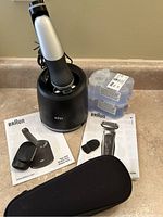 Braun electric shaver in charging and cleaning stand, two cleaning fluid cartridges, user manuals, small cleaning brush, and trimmer attachment arranged on countertop.