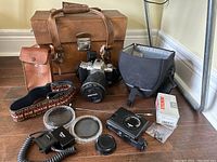 Full lot contents showing Pentax MZ-5N camera with zoom lens, flash, two filters in cases, camera battery, retro Nikon branded leather strap, soft camera case, large brown hard travel case with strap and metal latch.