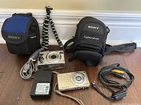 Two Sony Cybershot digital cameras alongside their cases, USB and AC cables, battery charger, extra battery, and small tripod on wooden floor.