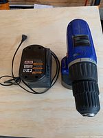 Blue cordless power drill with black chuck and speed dial, paired with black power corded battery charger on wood background.